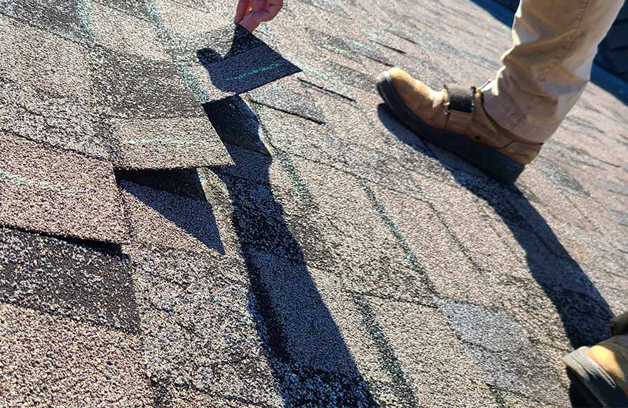 A person's hand lifting a damaged asphalt shingle on a roof doing a roof inspection for storm damage repair in Minneapolis, MN