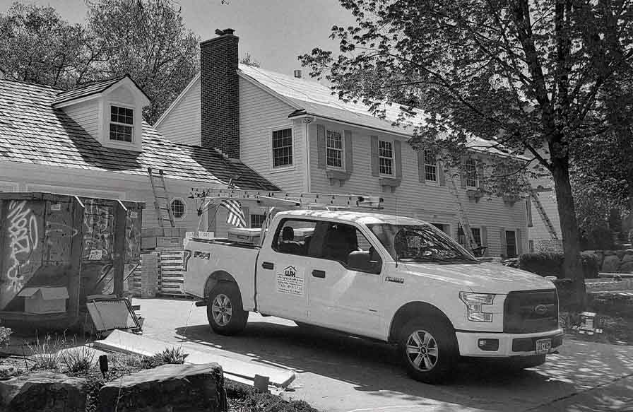 black and white image of a house undergoing roof repair in minneapolis. A white Wright at Home Roofing & Exteriors pickup truck with ladders on top is parked in front.