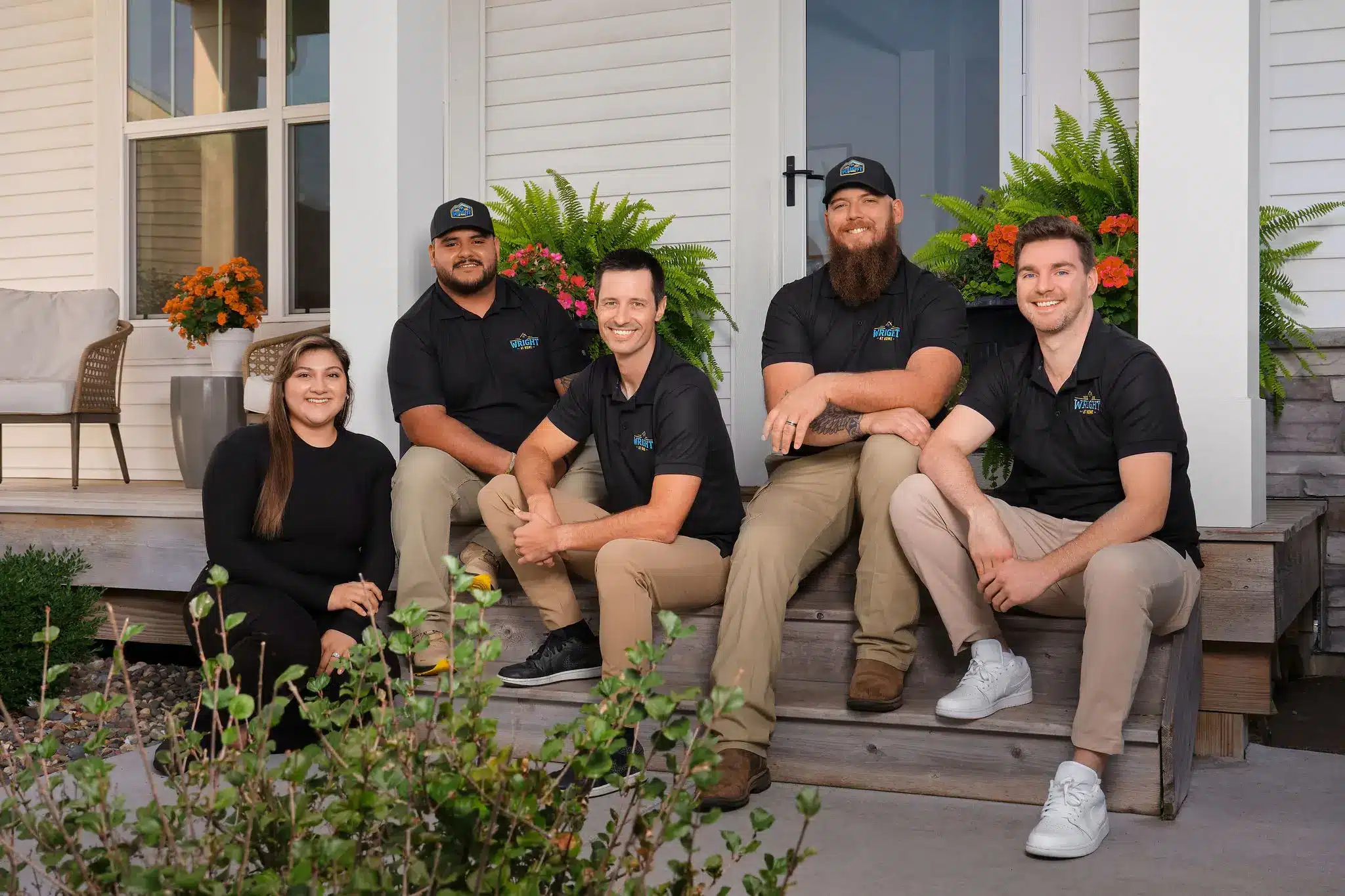 Five members of the Wright at Home Roofing & Exteriors team, wearing branded shirts, smiling and sitting together on the steps of a home’s front porch