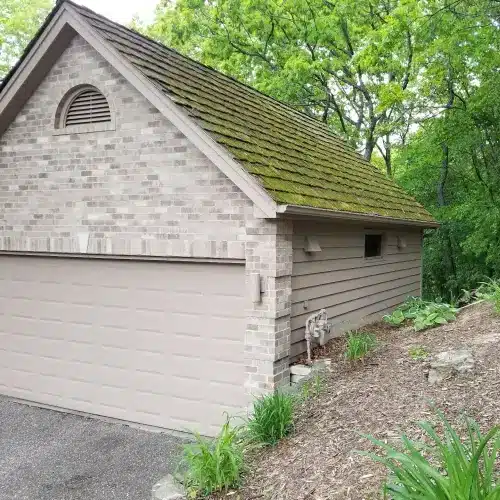 Minneapolis MN garage with moss-covered, weathered shingle roof, surrounded by greenery, demonstrating early stages of hail damage roof Minnesota by Wright at Home Roofing & Exteriors