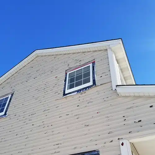 Severe hail damage on home siding and roof around window, clear blue sky, demonstrating hail damage roof Minnesota by Wright at Home Roofing & Exteriors in Minneapolis, MN